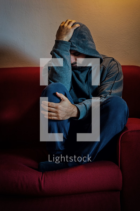 Young man is sitting alone on a red couch, holding his head in his hands, suffering from depression