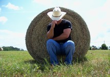 A cowboy with a cowboy hat tilts his hat to protect his face from the sun while resting in the shade behind a bale of hay in the country meadows of Florida.