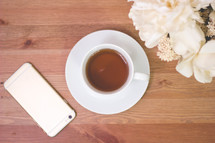 iPhone, coffee cup and saucer, and flowers on a wood table 