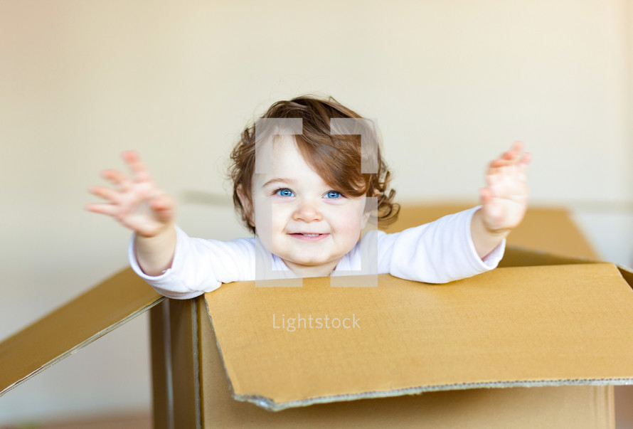 Cute smiling toddler baby girl sitting inside brown cardboard box.