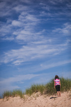 A little girl standing on a sand dune looking at a blue sky.