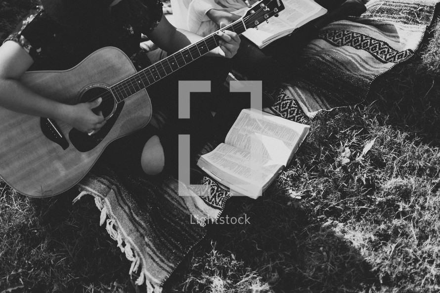 young woman sitting on a blanket playing a guitar and reading a Bible 