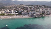 Aerial view of Waterfront Himare Buildings by turquoise colored water, Mountains in Background, Albania 