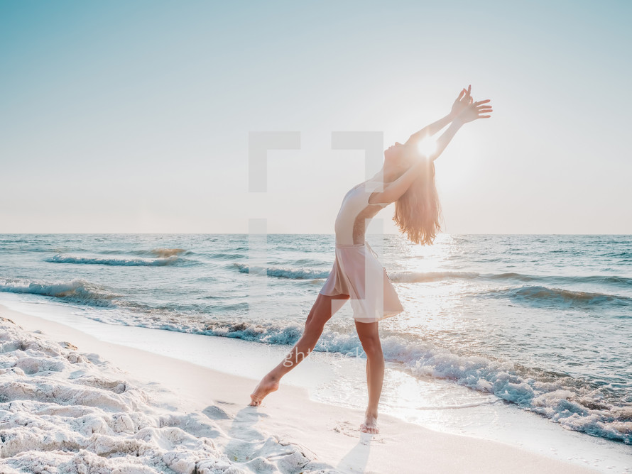 Slender beautiful ballerina in white dress dancing ballet on sea or ocean sandy beach in morning light. Concept of art, nature beauty.
