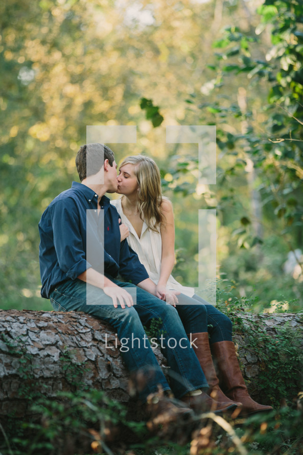 man and woman kissing while sitting on the trunk of a fallen tree