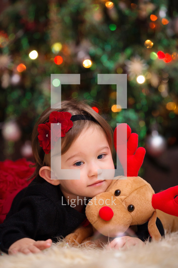 toddler girl on a fur rug with a stuffed animal reindeer under a Christmas tree