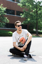 Smiling man wearing sunglasses sitting outside on a skateboard.
