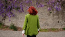 Back of a woman walking towards a cement wall covered with wisteria.