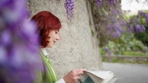 Woman reading a newspaper surrounded by wisteria.