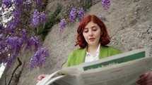 Woman reading a newspaper under wisteria.