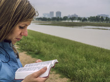 A woman reading the Bible near a river.