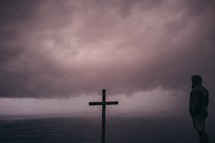 Wooden cross and a man on the ocean water, covered by low-lying storm clouds.