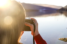 a girl taking a picture with a camera by a lake 