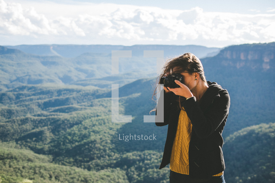 woman standing on a mountain top under a cloudy sky taking pictures with a camera 