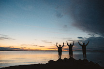silhouettes of men with raised hands standing by a lake at sunset 