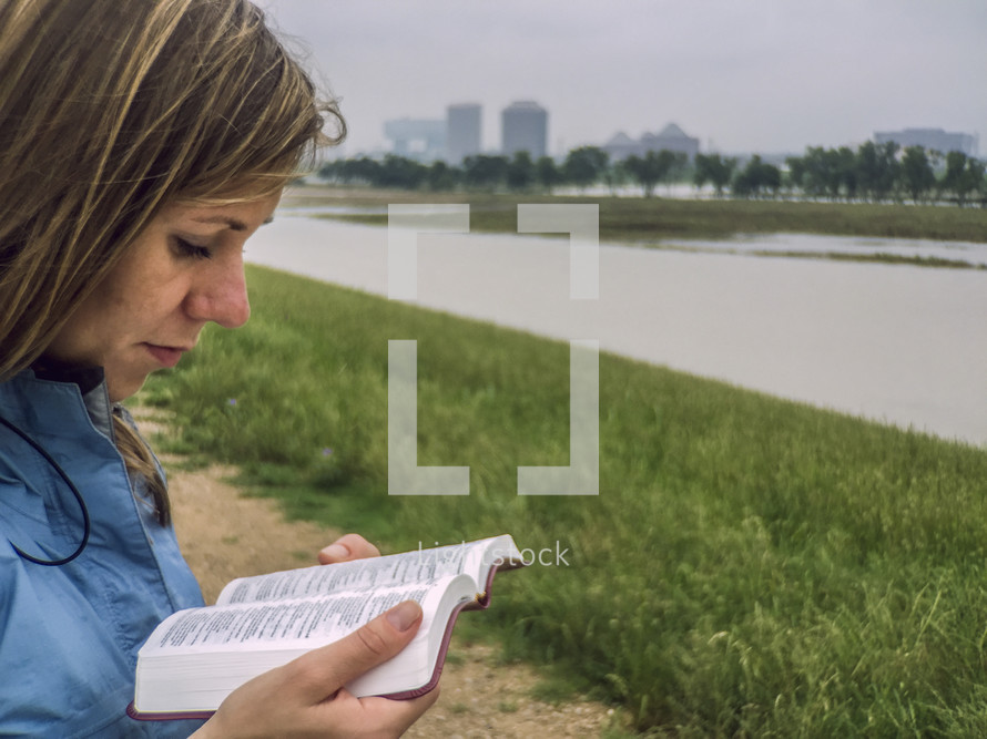 A woman reading the Bible near a river.