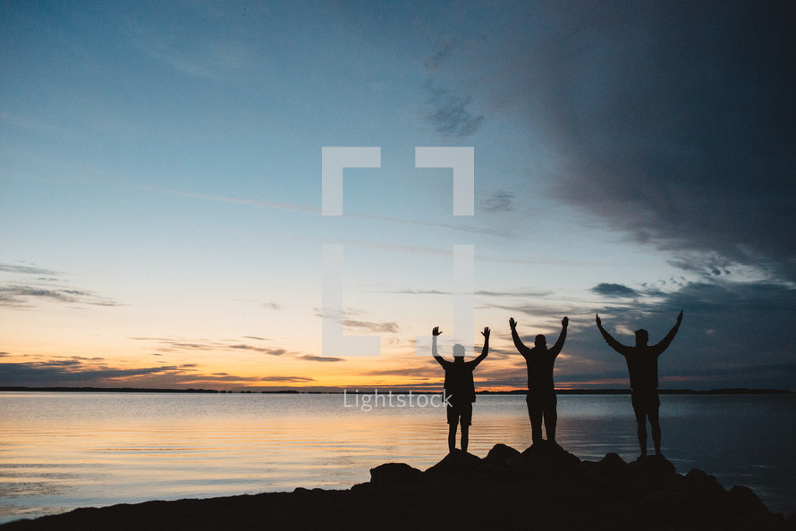 silhouettes of men with raised hands standing by a lake at sunset 