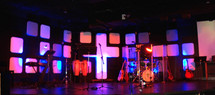 A lighted sound stage for recording music, playing concerts or having a live worship band. The stage is decorated with a cross, lighting, lighted panels and musical instruments including guitars, drums, microphones, bass guitar and other instruments for leading a live praise and worship church service. 