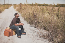 man sitting on luggage on a dirt road praying 