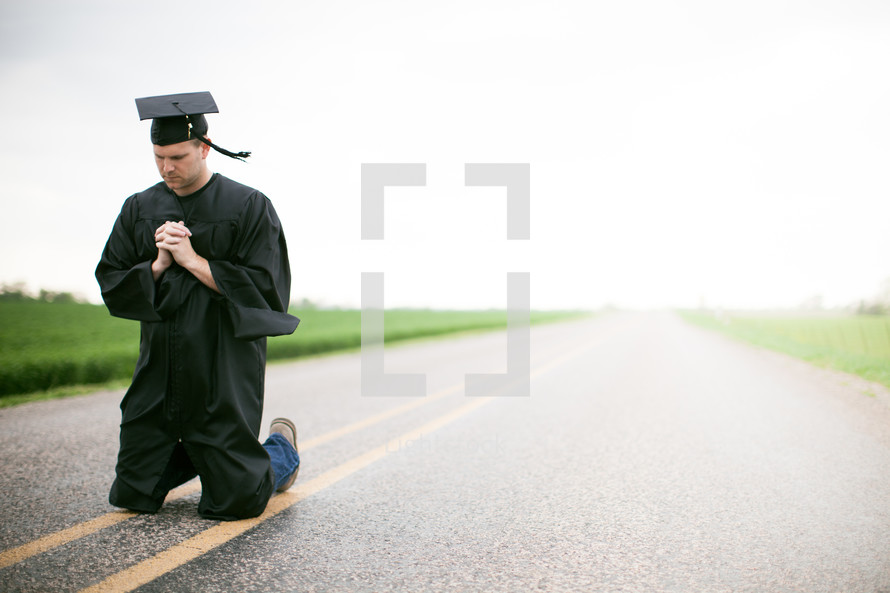 Graduate kneeling in the middle of the road in prayer.