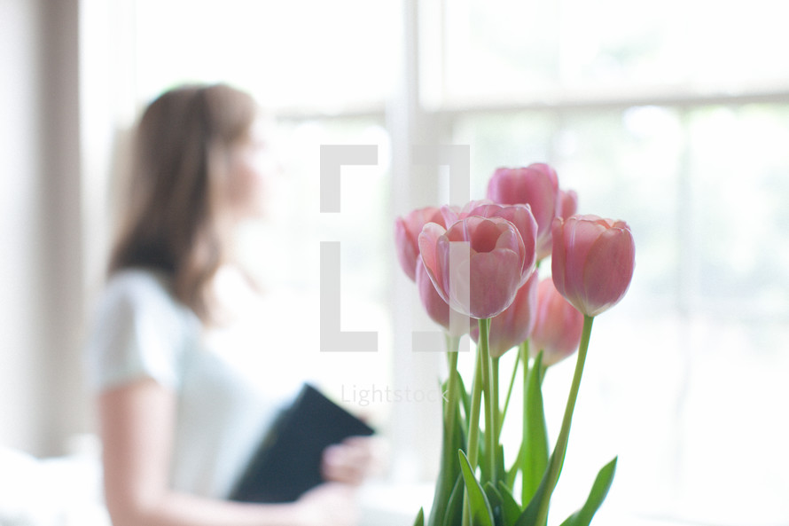 woman holding a Bible looking out a window and pink tulips