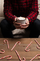 a man holding a plaid shirt holding hot cocoa and candy canes spread out on a table 