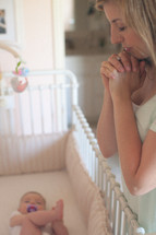 mother praying next to baby in a crib