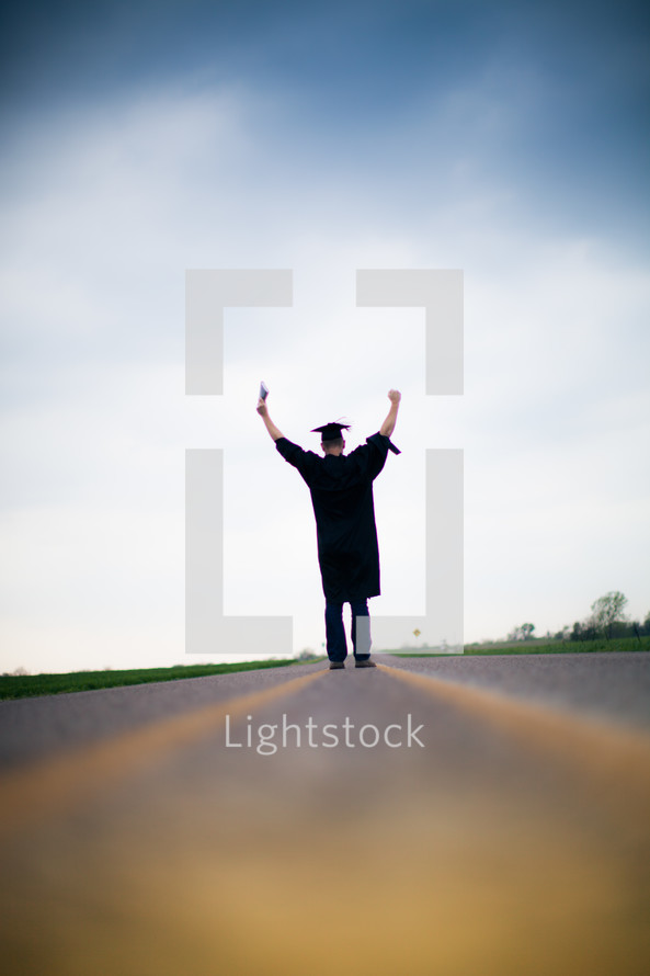 Graduate holding Bible walking down the middle of the road with arms raised.