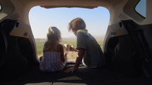 Brother and sister drinking from mugs in the back of a car overlooking a farm