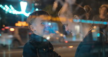 Son and mother in the busy street at night. Excited child talking to the mom, she embracing and kissing him. View through the glass