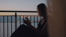 Woman sitting on a balcony at dusk using a smart tablet.