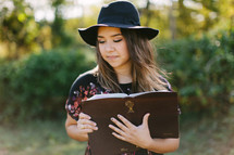portrait of a teen girl in a hat standing outdoors reading a Bible 
