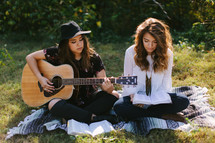 teen girls sitting on a blanket outdoors playing a guitar and singing 