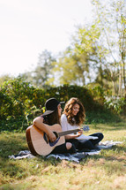 young women sitting on a blanket outdoors playing a guitar and singing 