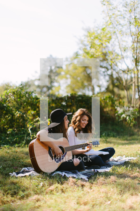 young women sitting on a blanket outdoors playing a guitar and singing 