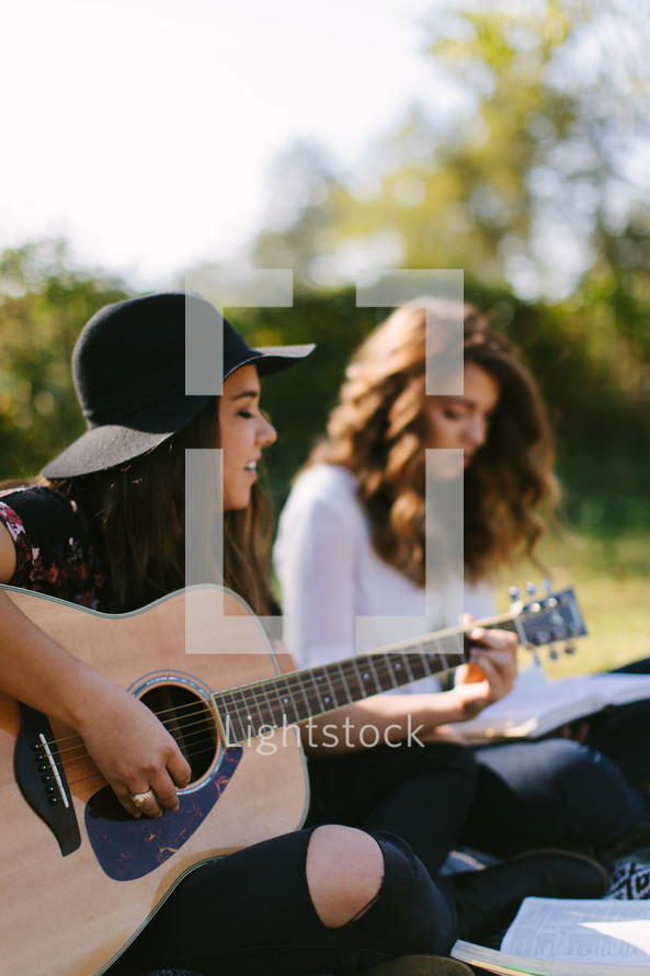 young women sitting on a blanket outdoors reading Bibles and playing guitars 