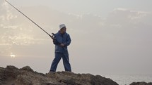 Old fisherman standing on sea side rocks and fishing against the sunset