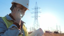 Engineer staying on field with telecommunications tower talking on phone and solving business issues. Man in protective helmet and special work clothes. Holds in hands plan for construction facility.