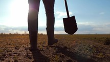 Legs of man agronomist close-up walking across field with shovel in his hands for plowing farm land. Agricultural production of natural healthy eat. Growing and selling large volumes of products.