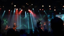 Silhouette of an audience with arms raised at a concert with colorful stage lighting.