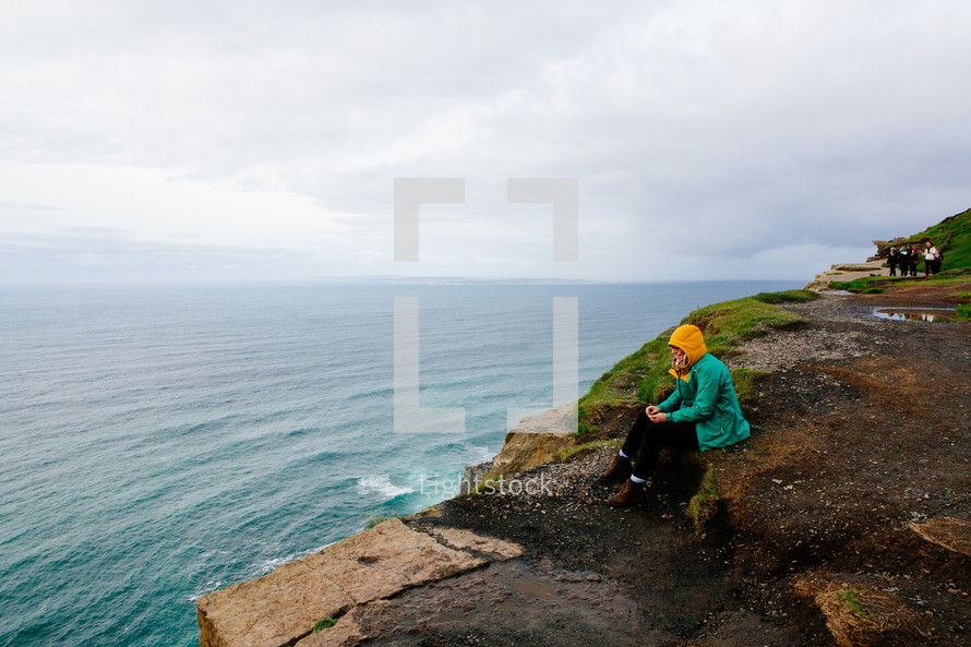 a woman in a raincoat sitting on a shore on the Cliffs of Moher 