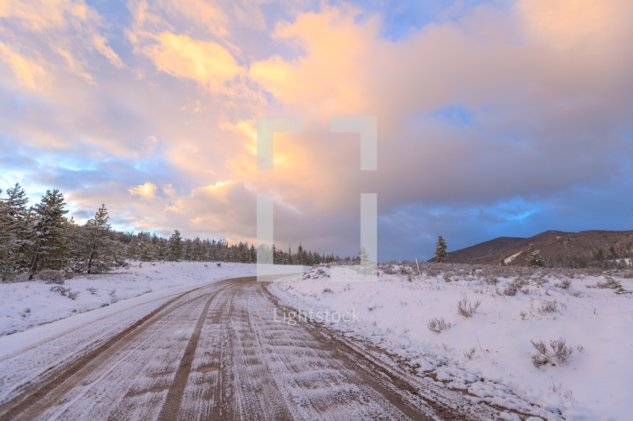 Vehicle tracks in snow on a dirt road near mountains and trees during winter with colorful clouds at sunset 