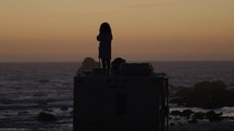 Woman stands on RV watching ocean at dusk, handheld