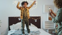 Kid Jumping in Bedroom Decorated for Christmas, Mom Taking Video with Phone
