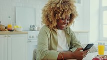 Smiling Black Woman Texting on Smartphone during Breakfast at Home
