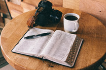 An open Bible, cup of coffee, and camera on a table.