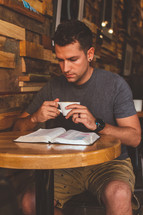 A man sits at a table in a cafe reading the Bible and drinking coffee.