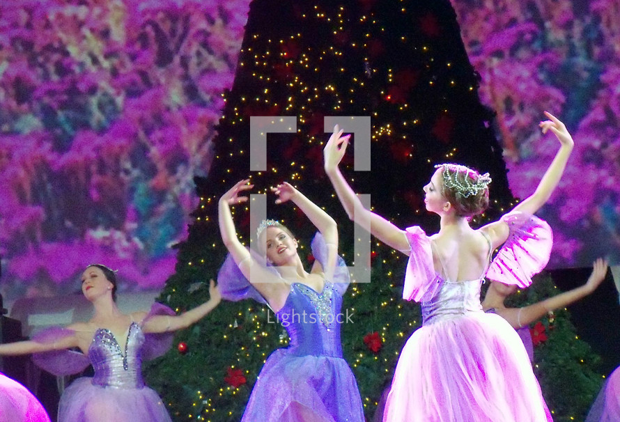 A group of Ballerinas dance in front of a Christmas tree on stage during a performance of The Nutcracker during a Christmas celebration surrounded by Christmas lights and purple and violet lighting. 