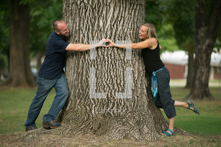 couple making a heart with their hands around a tree trunk