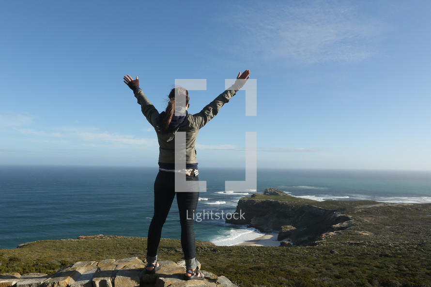 woman standing on a mountain top over the ocean with arms raised in praise and worship